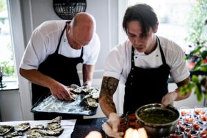 Two chefs in a restaurant kitchen preparing gourmet dishes with fresh ingredients and focused expressions.